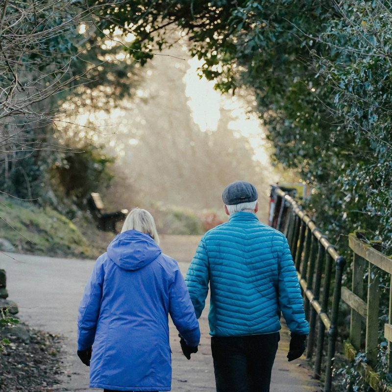 An older couple walking together at golden hour, in blue jackets — Walk Past Sixty, the senior edition