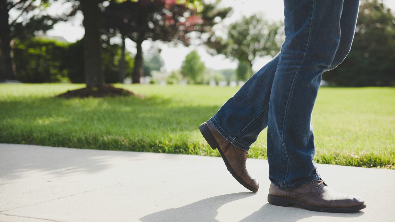 A walker in brown shoes and blue jeans striding along a concrete path beside a strip of green grass.